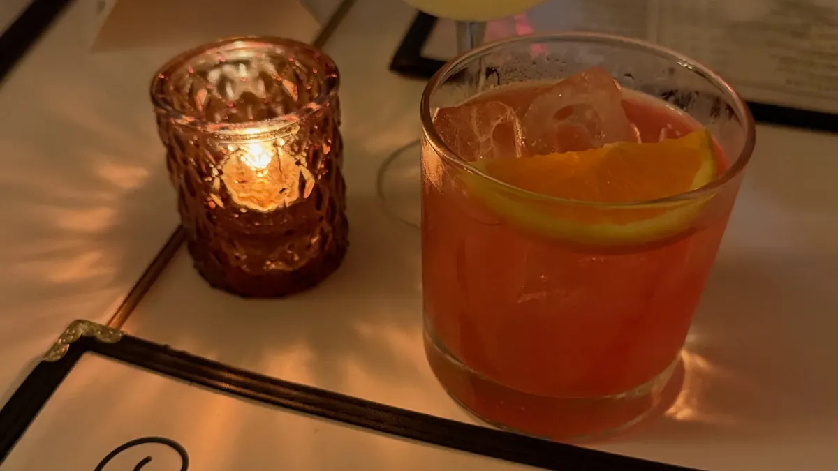 Two cocktails, one with an orange slice, beside a flickering candle and a restaurant menu on a candlelit table.