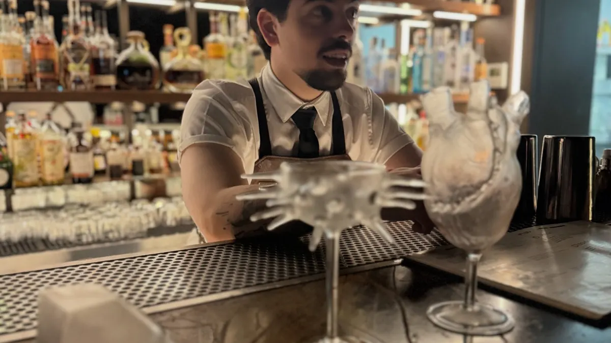 A smiling bartender in a vest and tie leaning on a polished bar, with bottles and glassware behind him.