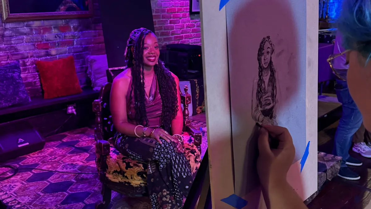 A live artist sketches a portrait of a woman posing on a lounge chair in a purple-lit studio.