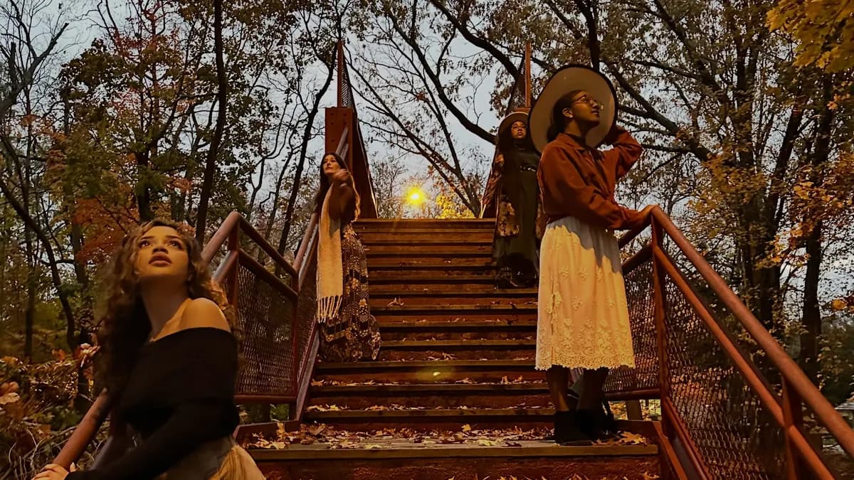 Women in long skirts, sweaters and wide-brimmed hats posing on a rustic wooden staircase surrounded by autumn trees.