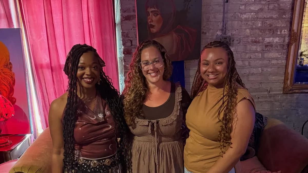 Cristina Vann smiling between two women in a brick-walled studio with a pink curtain and a painted portrait behind them.