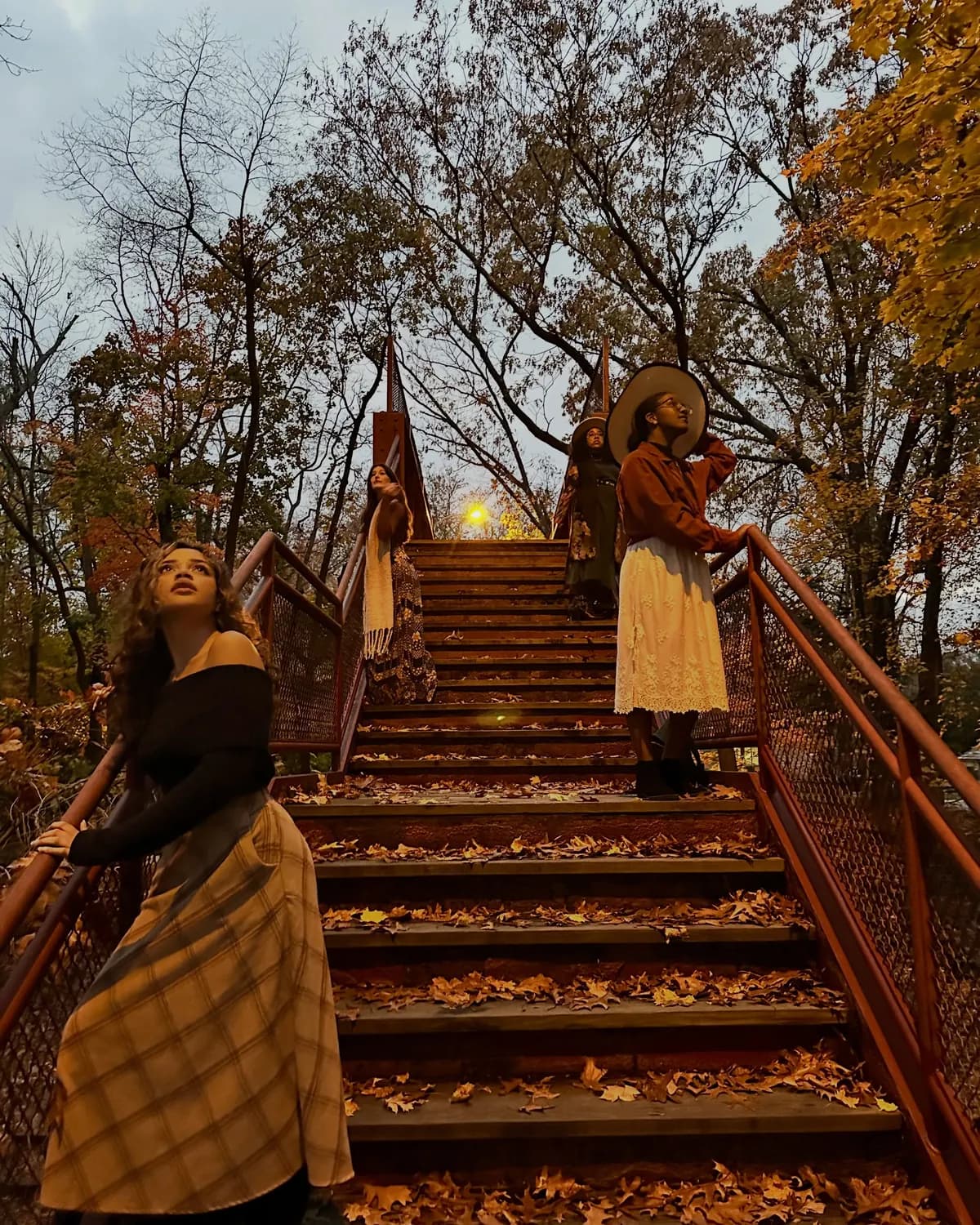 Women in long skirts and hats pose on a rustic wooden staircase among fall trees.